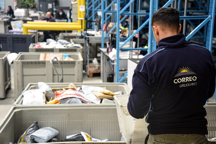 El Correo Uruguayo alerta a la población sobre intentos de fraude que suplantan su identidad Persona con una campera del Correo Uruguayo, de espaldas, trabaja con cajas con objetos a su lado